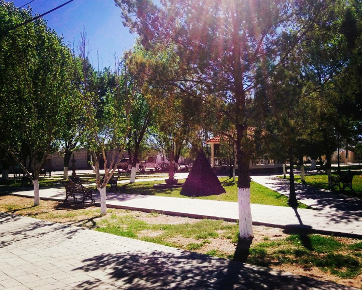 Plaza arbolada con bancas de hierro y un kiosco de teja roja al fondo